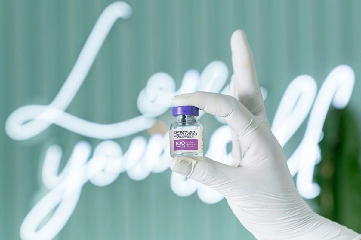 Injector Holding a vial of Botox in front of a neon sign that says Love yourself