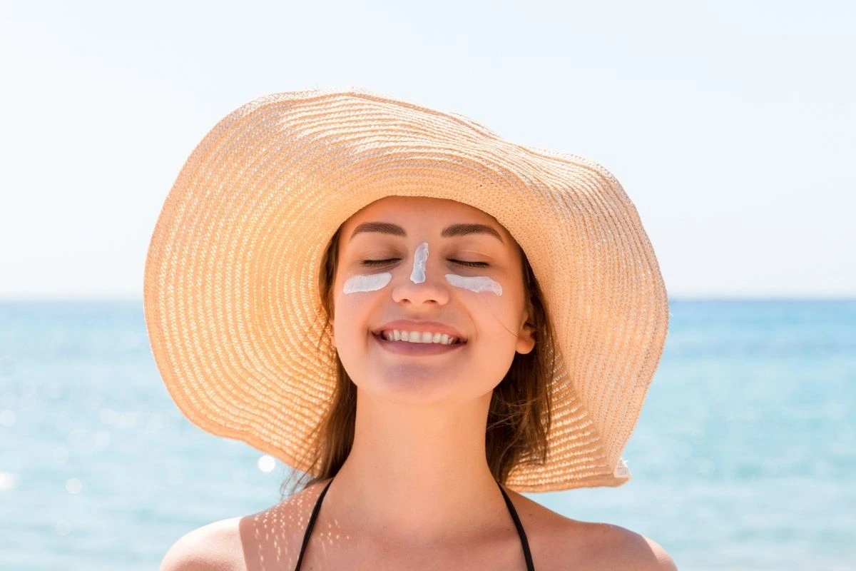 Woman in hat at the beach putting sunscreen on her cheeks and nose.
